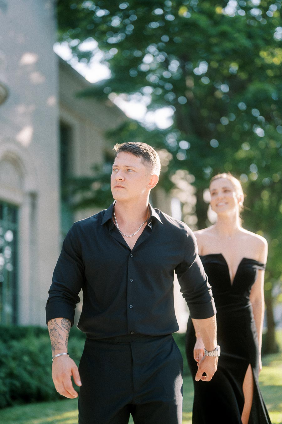 Couple in black formalwear walking outdoors beside a building in warm late-afternoon light