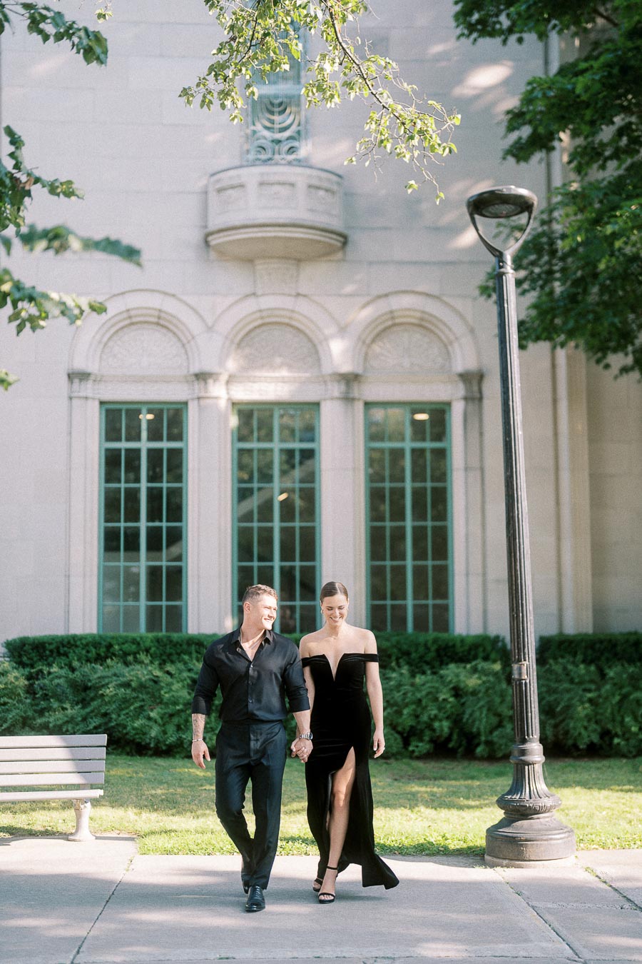 Couple holding hands while walking past arched windows, greenery, a bench, and a lamppost in Toronto.