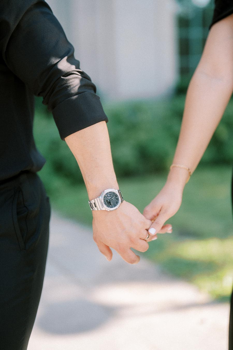 Couple holding hands outdoors, with engagement ring and silver watch visible in soft sunlight.