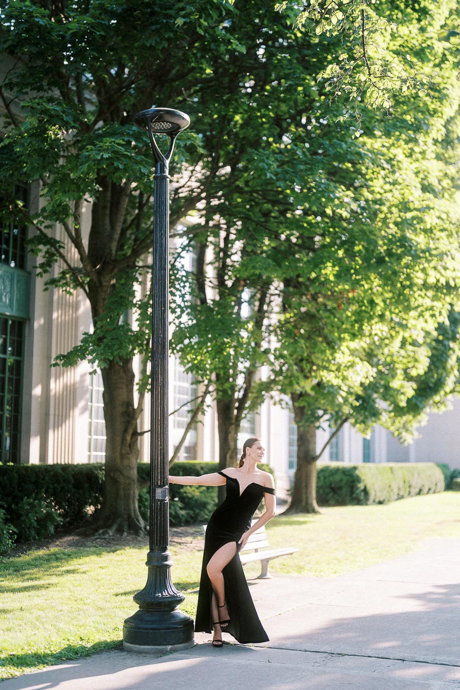 Woman in a black gown poses beside a lamppost on a sunlit tree-lined Toronto walkway.