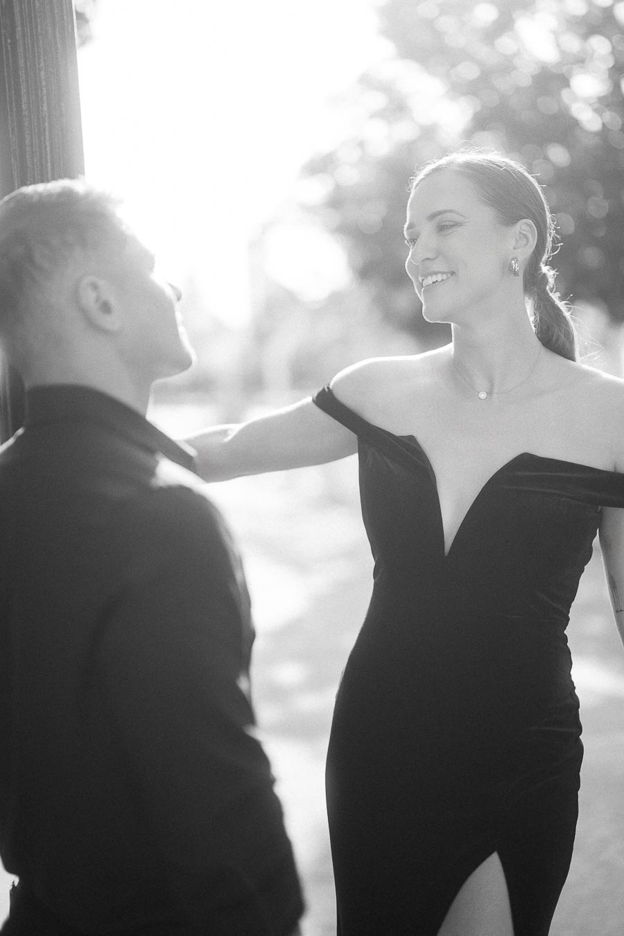 Woman in off-shoulder dress smiles at her partner in soft black-and-white backlight outdoors.