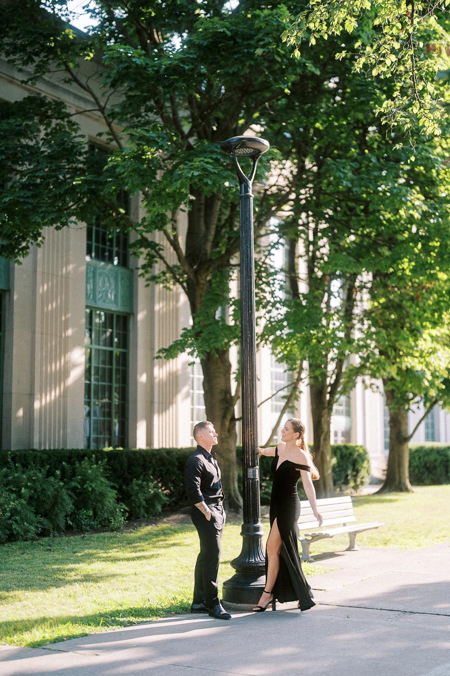 Engaged couple in black formalwear posing beside a lamppost on a leafy Toronto walkway