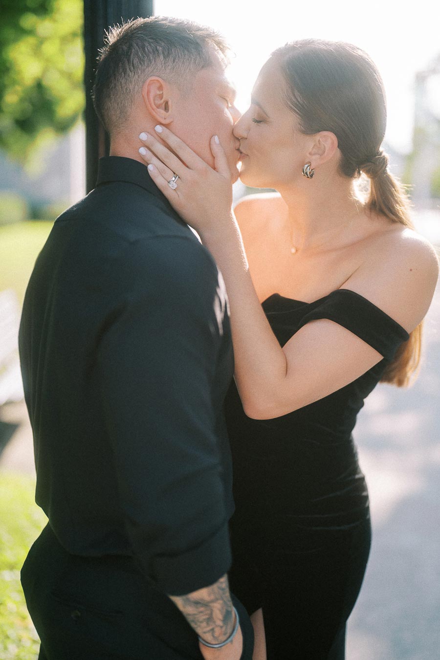Couple in black outfits kissing outdoors as the bride-to-be shows her engagement ring.