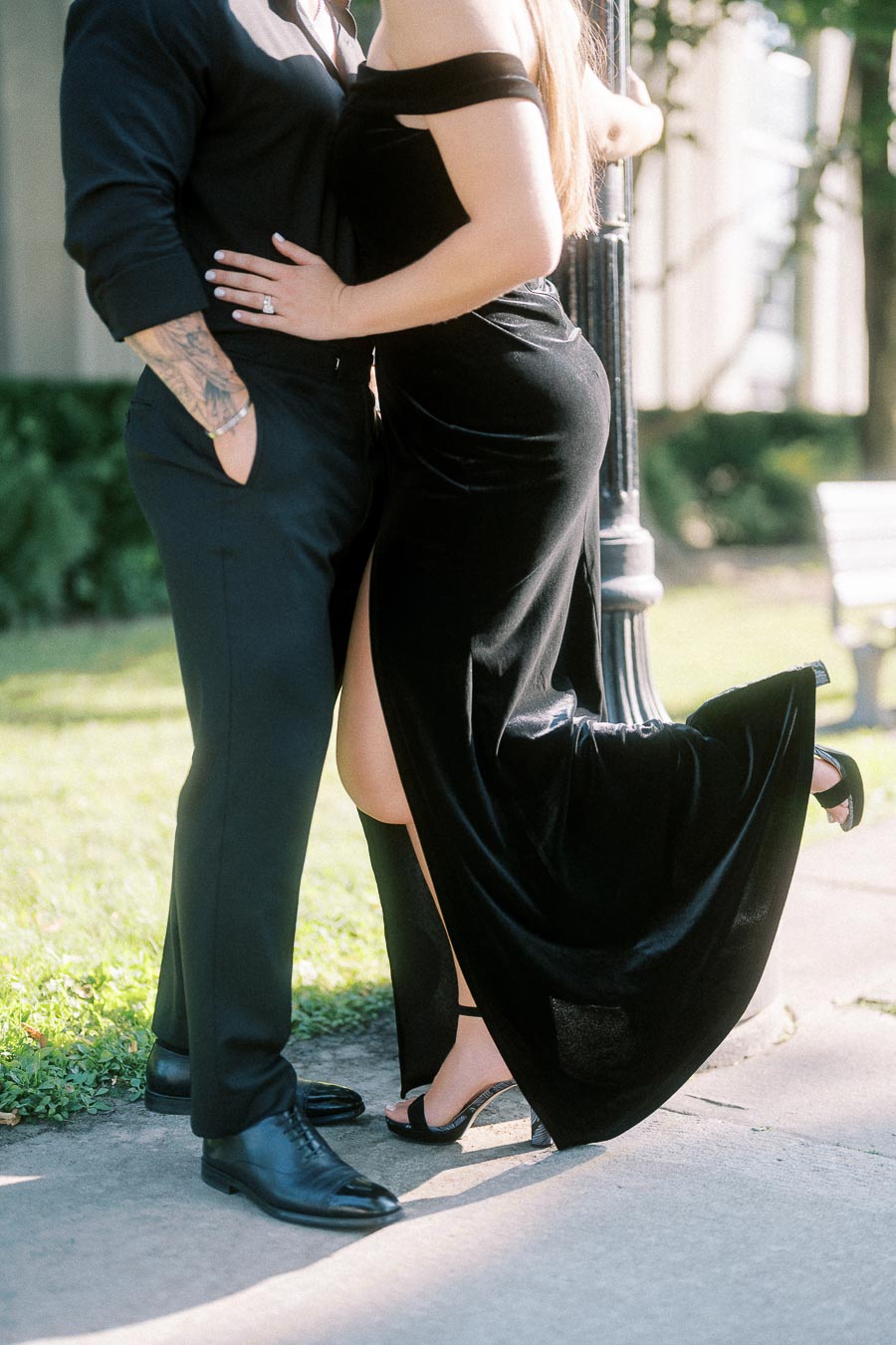 Couple in black formal outfits embraces on a sunny Toronto walkway, her engagement ring visible.