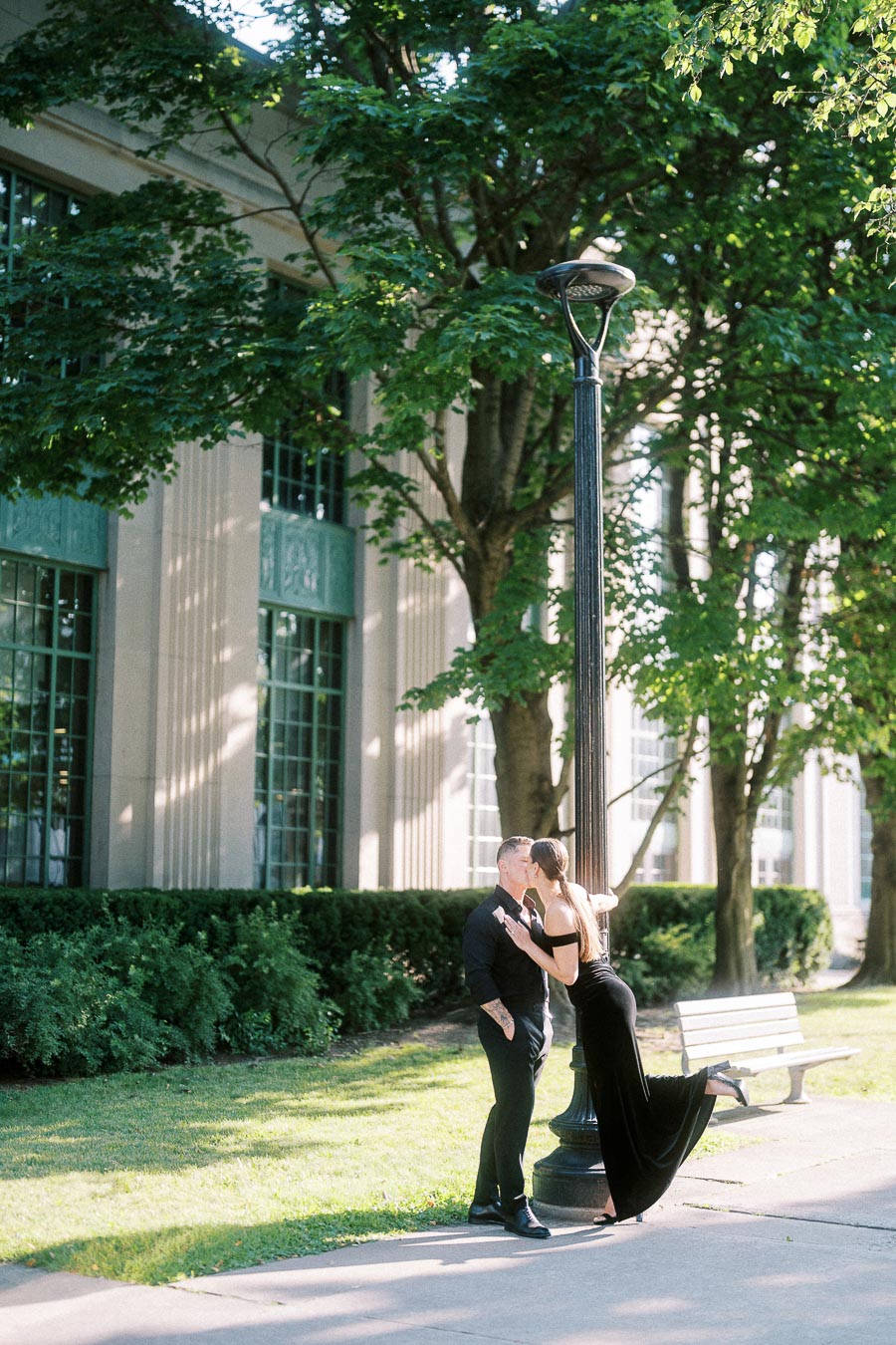 Couple in black outfits kissing beside a lamppost on a leafy Toronto walkway.