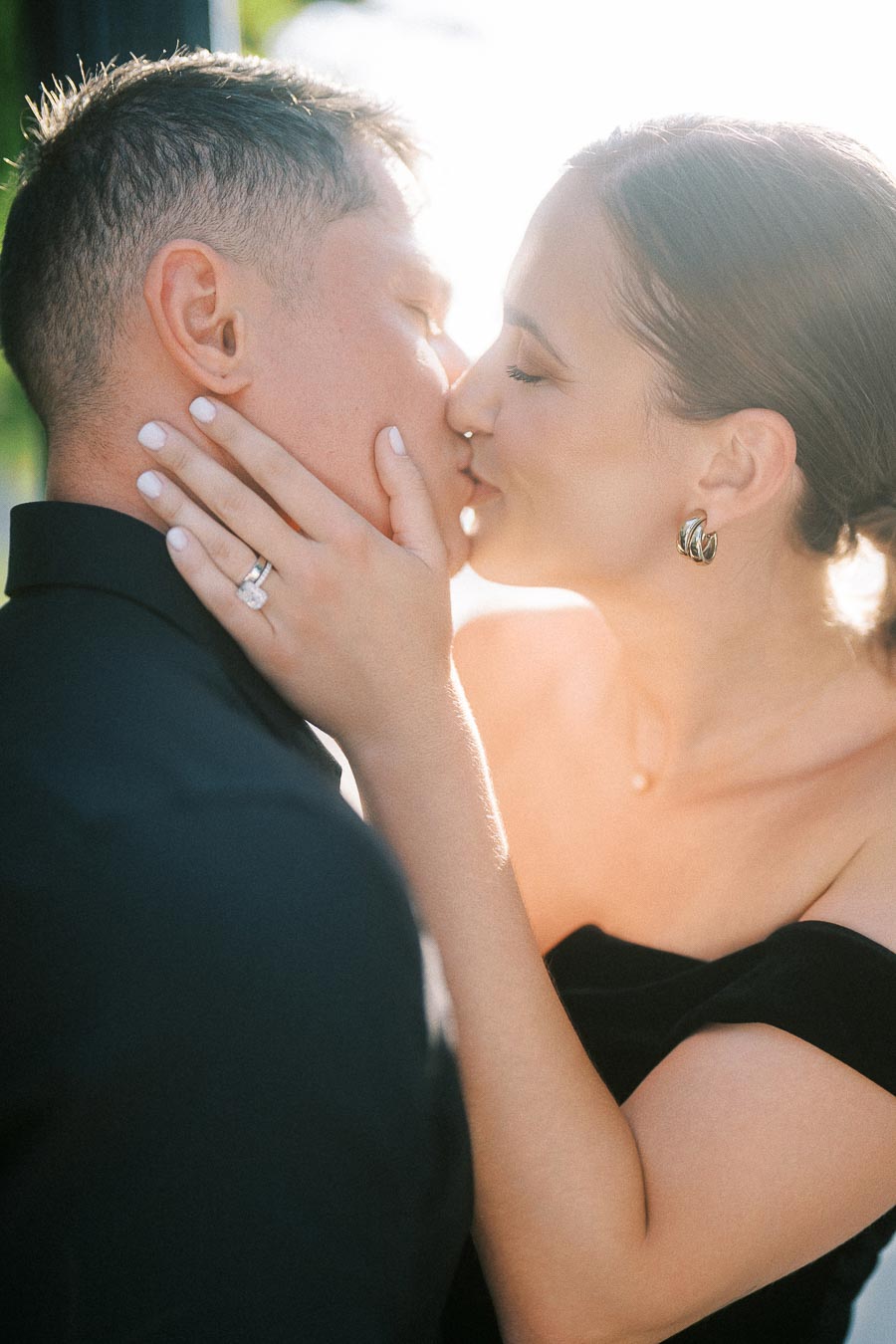 Couple kissing closely in warm backlight, her hand showing engagement ring on his cheek.
