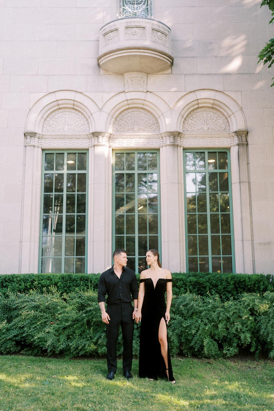 Engaged couple in formal black attire standing on grass before tall arched windows and greenery.