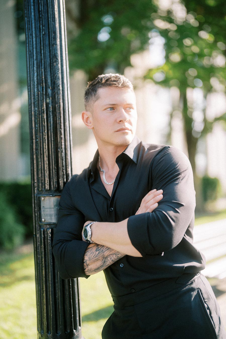 Groom in black shirt leans against a lamppost with arms crossed in soft outdoor light.