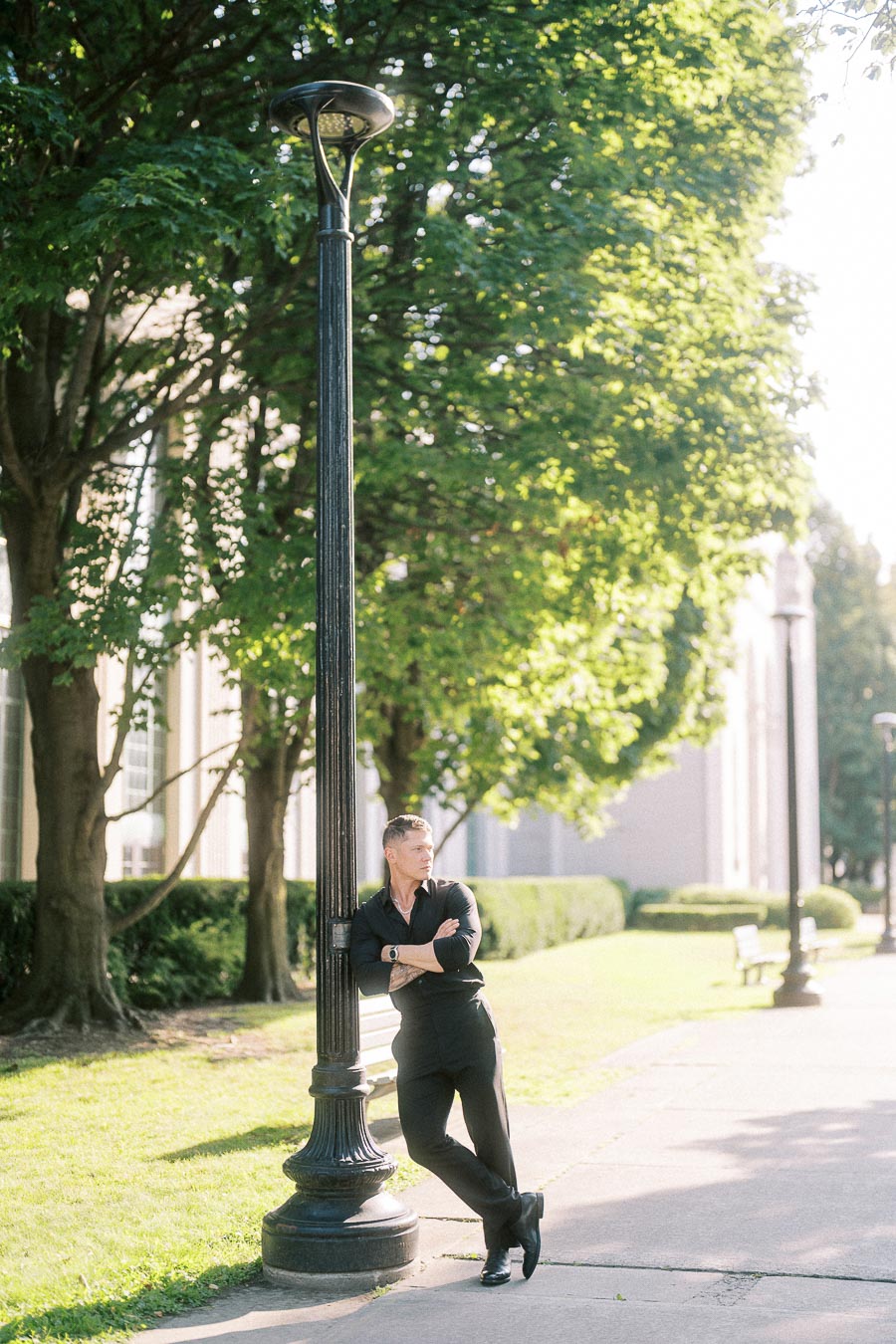 Man in black leaning against a lamppost on a sunlit tree-lined Toronto path.