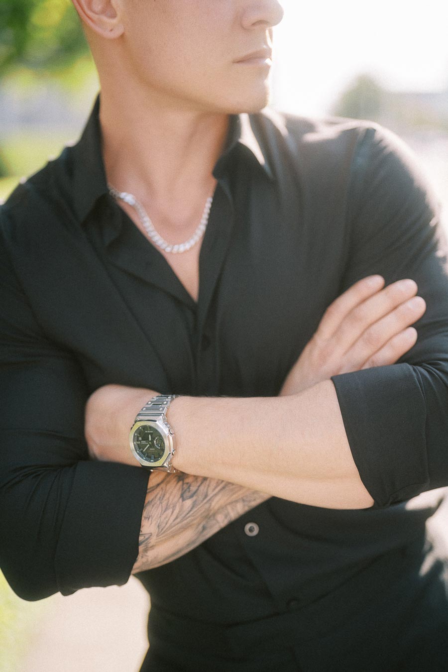 Man in black shirt with crossed arms, silver watch, chain necklace, and soft outdoor light.