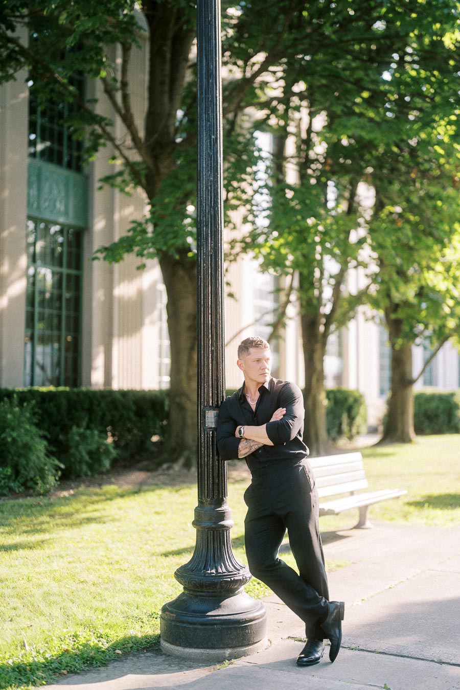 Man in black outfit leans against a lamppost on a sunlit tree-lined Toronto walkway.