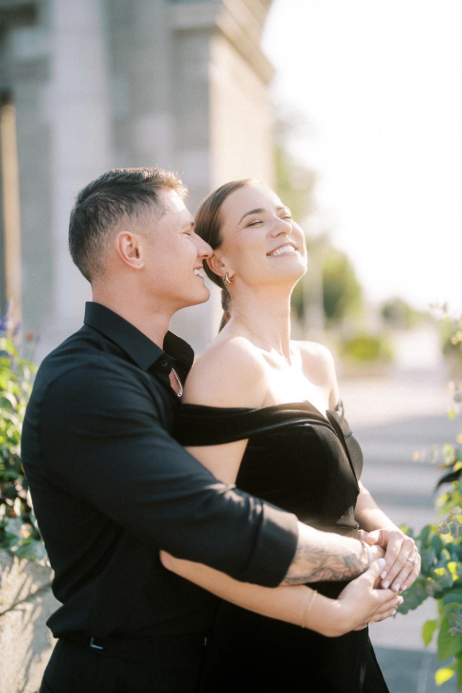 Smiling couple in black outfits embracing outdoors in warm sunlight along a Toronto streetscape.