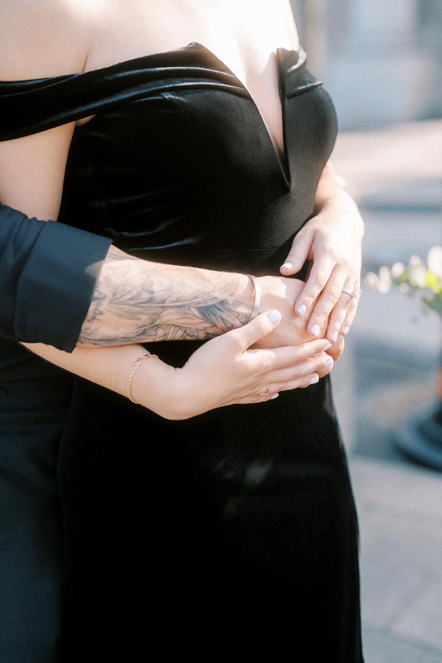 Close-up of a couple embracing, hands resting over a black dress with an engagement ring visible.