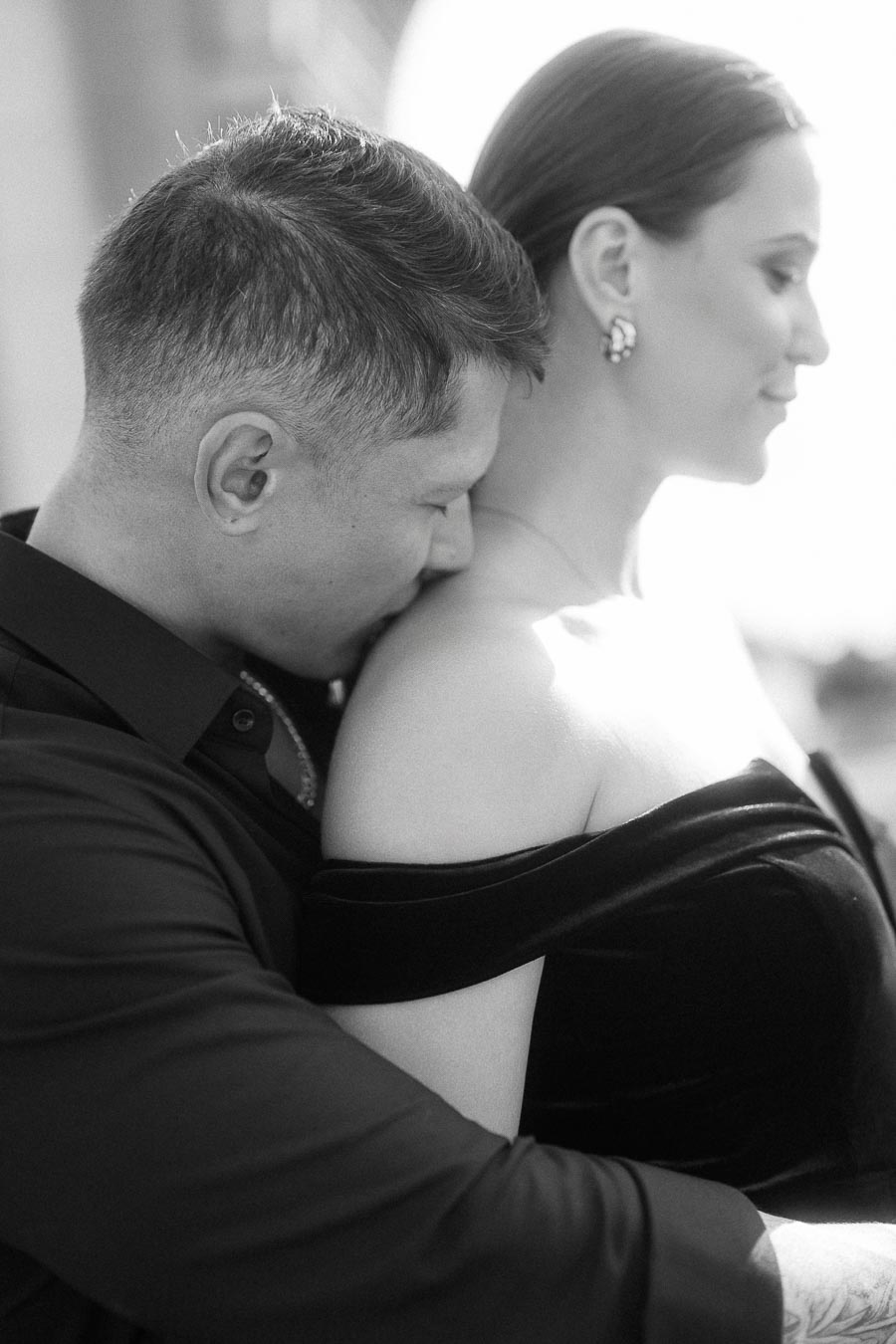 Black-and-white close portrait of a couple embracing, with the man resting his face on her shoulder.