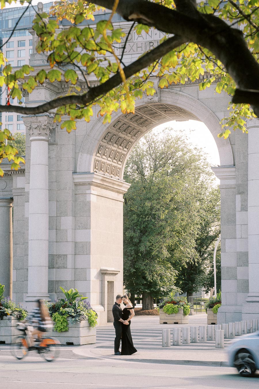 Couple embraces beneath the Princes’ Gates arch at Exhibition Place with passing cyclist and car.