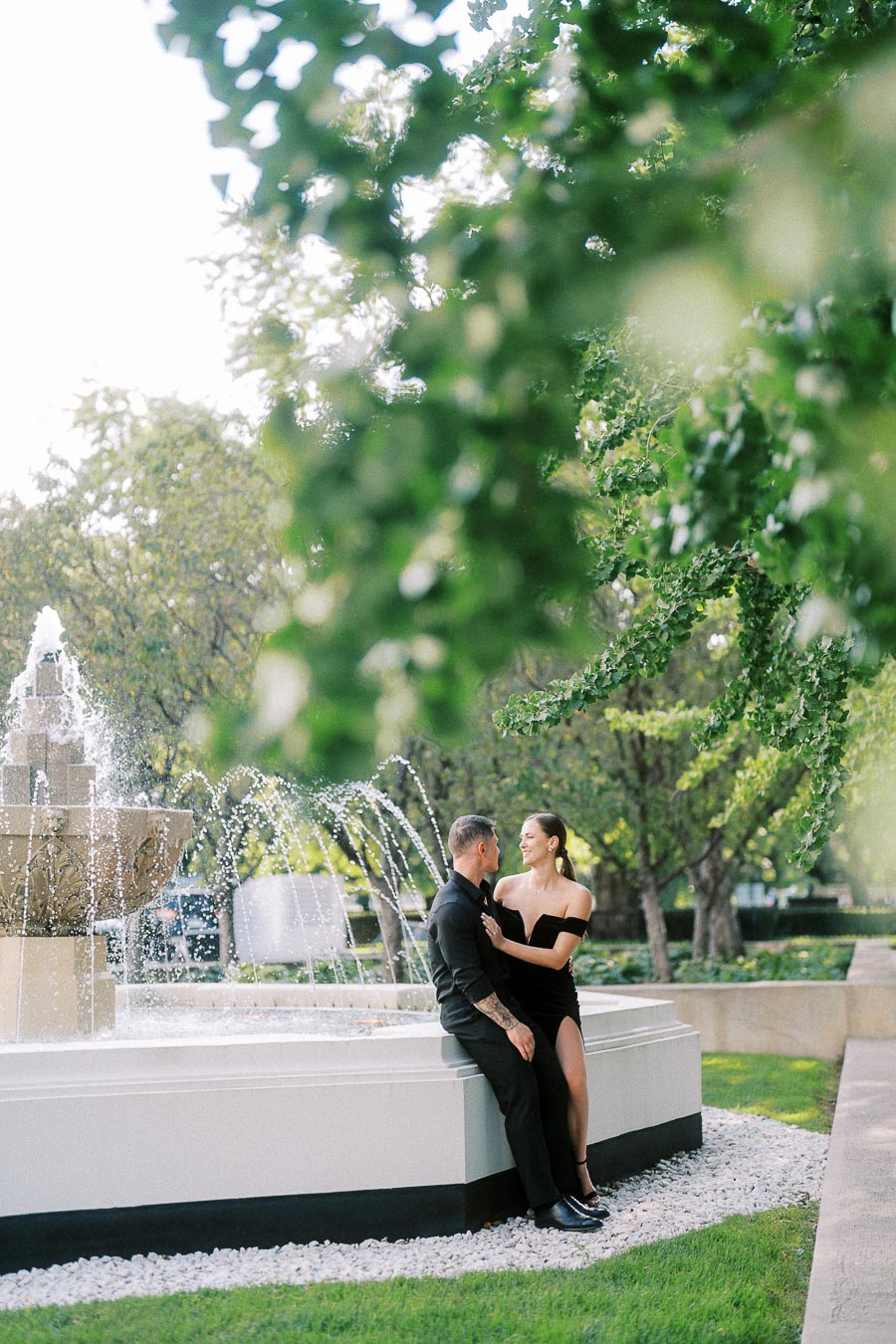 Couple in black outfits sitting beside a garden fountain under leafy trees in Toronto.