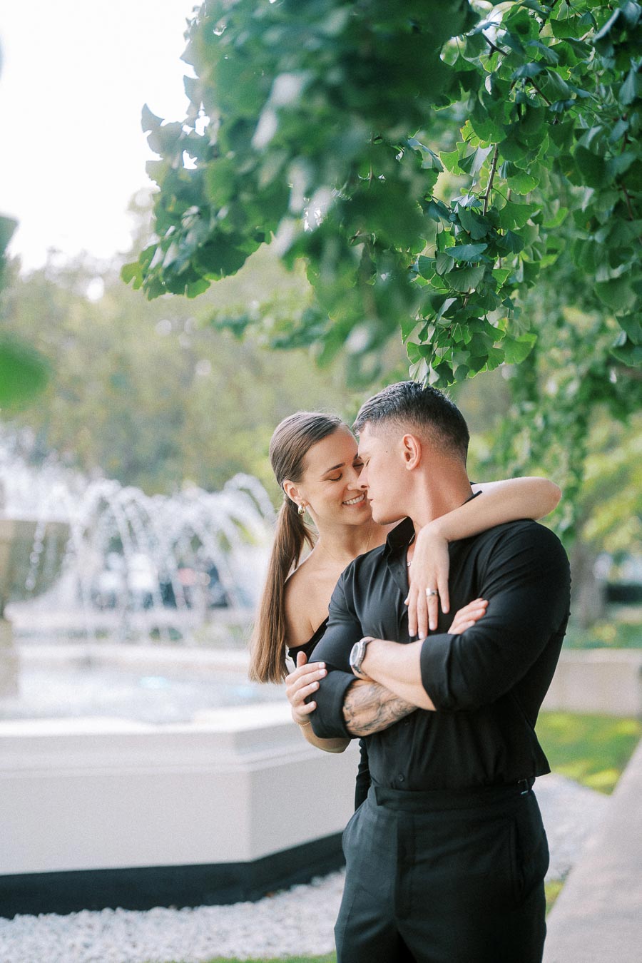 Couple embracing beneath leafy branches beside a fountain, with her ring visible on his shoulder.