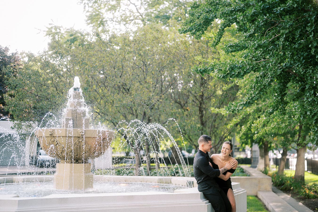 Couple laughing together beside a fountain in a leafy Toronto garden setting.