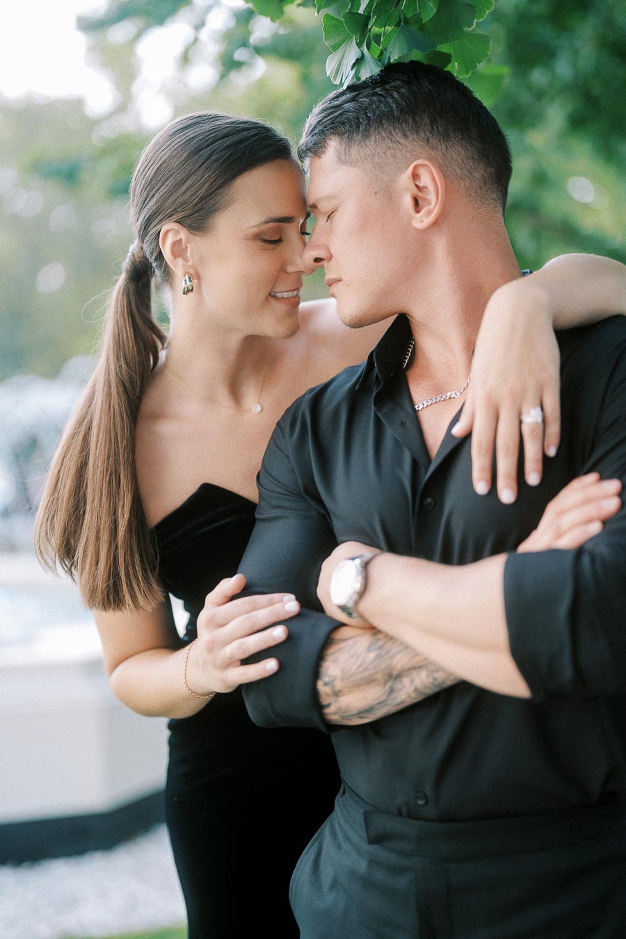 Couple in black outfits share a close embrace under leafy greenery, showing her engagement ring.