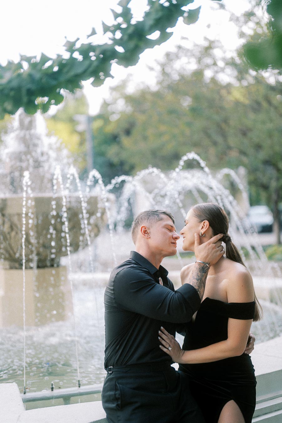 Couple in black outfits embracing beside a fountain with arcing water jets and leafy greenery.