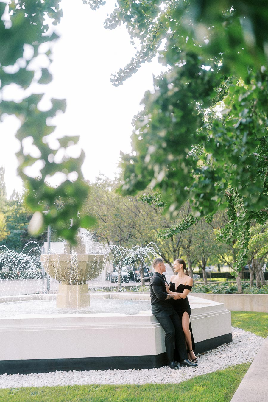Couple in black formal outfits sitting beside a garden fountain under leafy trees in Toronto.