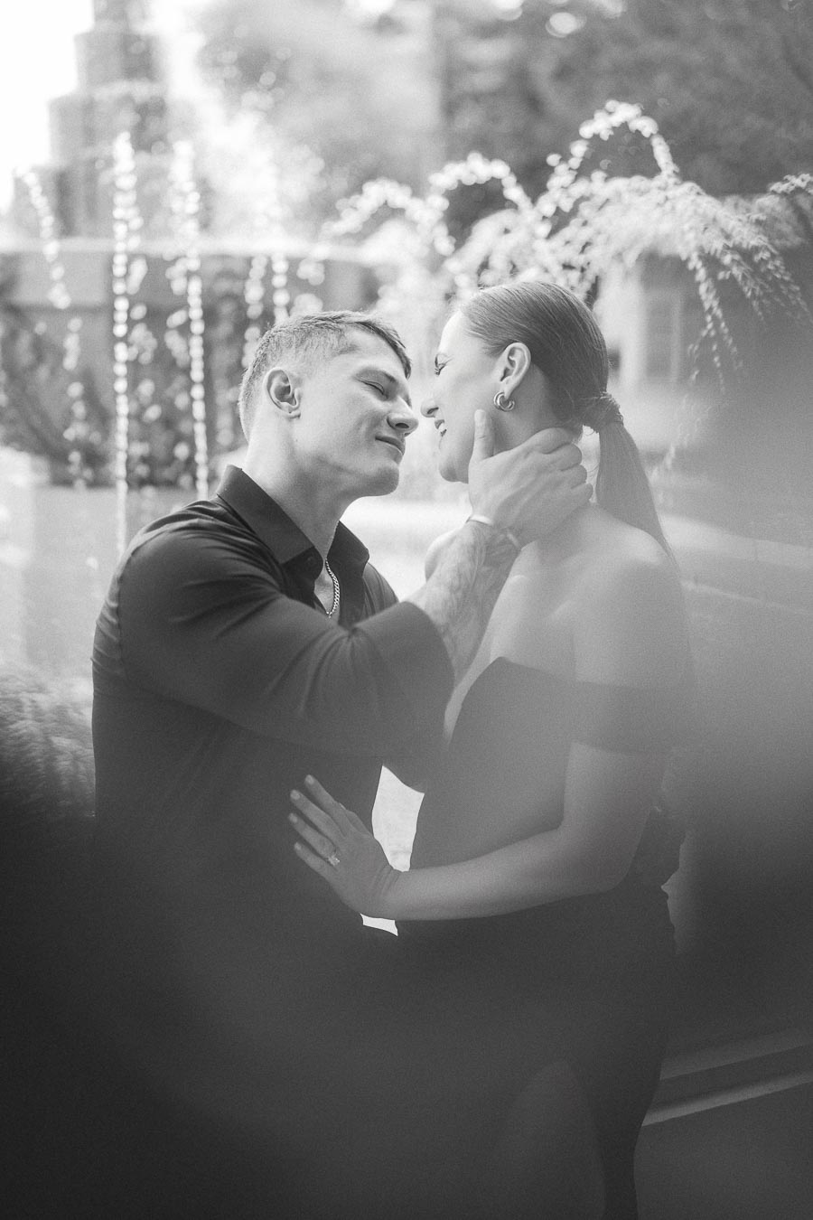 Black and white portrait of an engaged couple smiling closely beside a fountain and garden foliage.