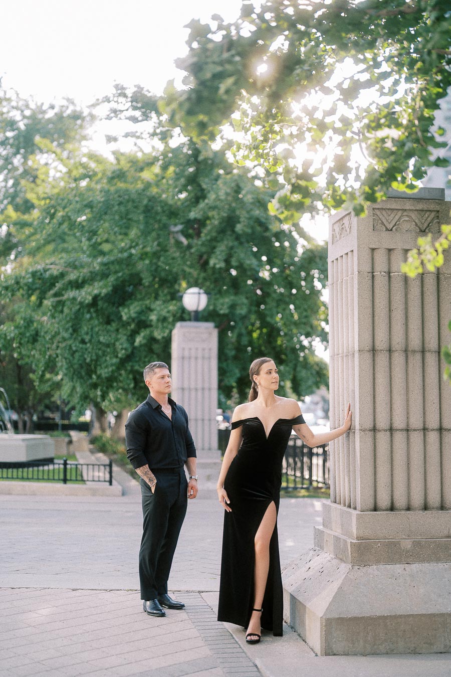 Couple in black formalwear poses beside stone pillars under leafy trees in Toronto.