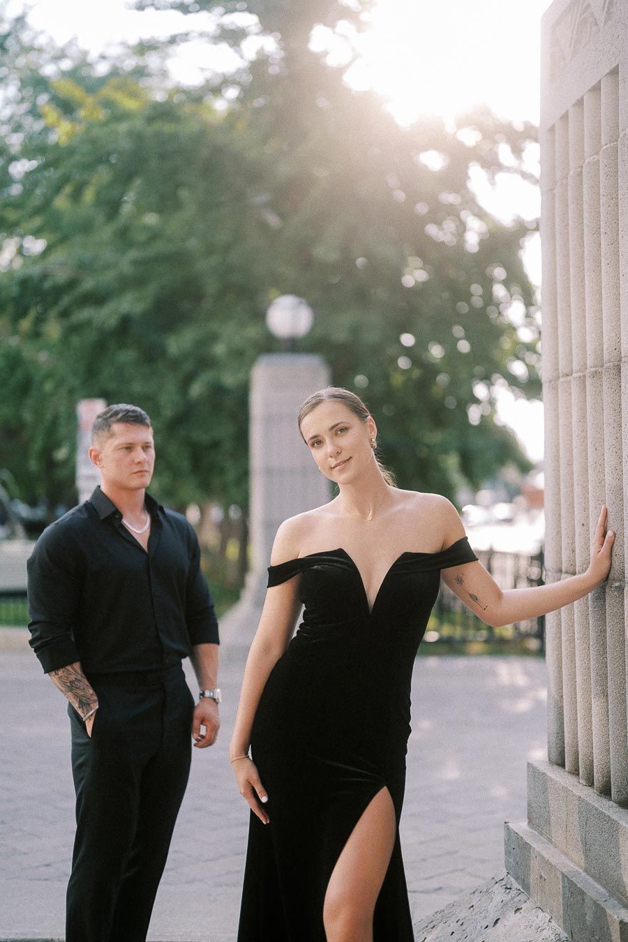 Couple in black attire posing beside stone columns with trees and warm evening light behind them.