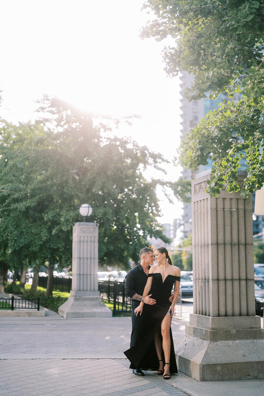 Couple in black formalwear embraces beside stone columns on a sunny downtown Toronto sidewalk.