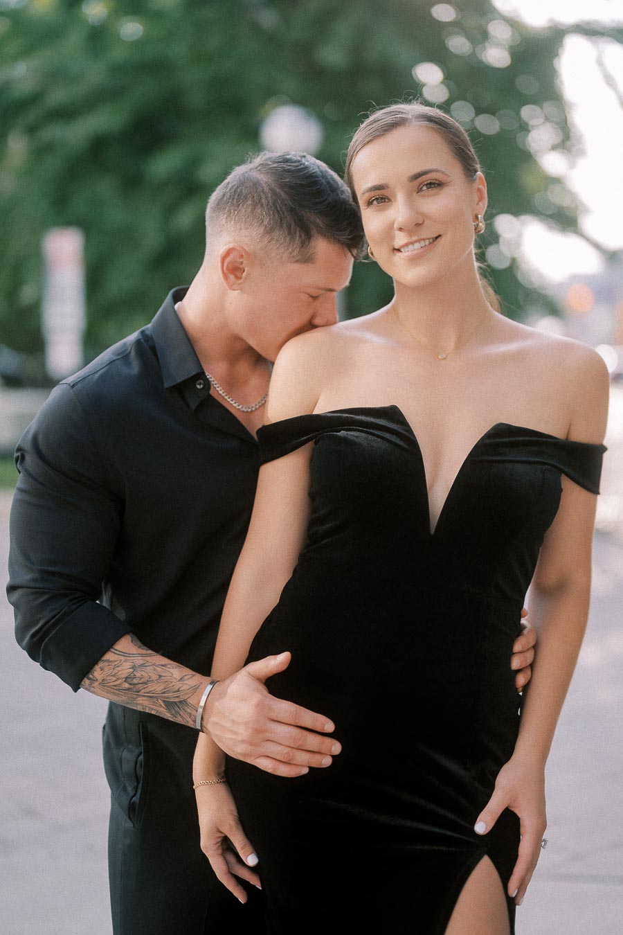 Couple in black formal outfits posing closely on a tree-lined Toronto sidewalk.