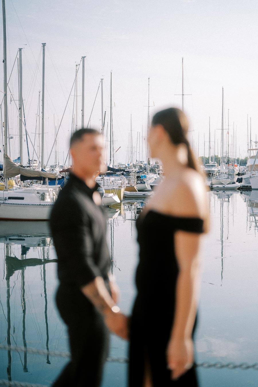 Soft-focused couple holding hands beside sailboats and calm marina water in Toronto.