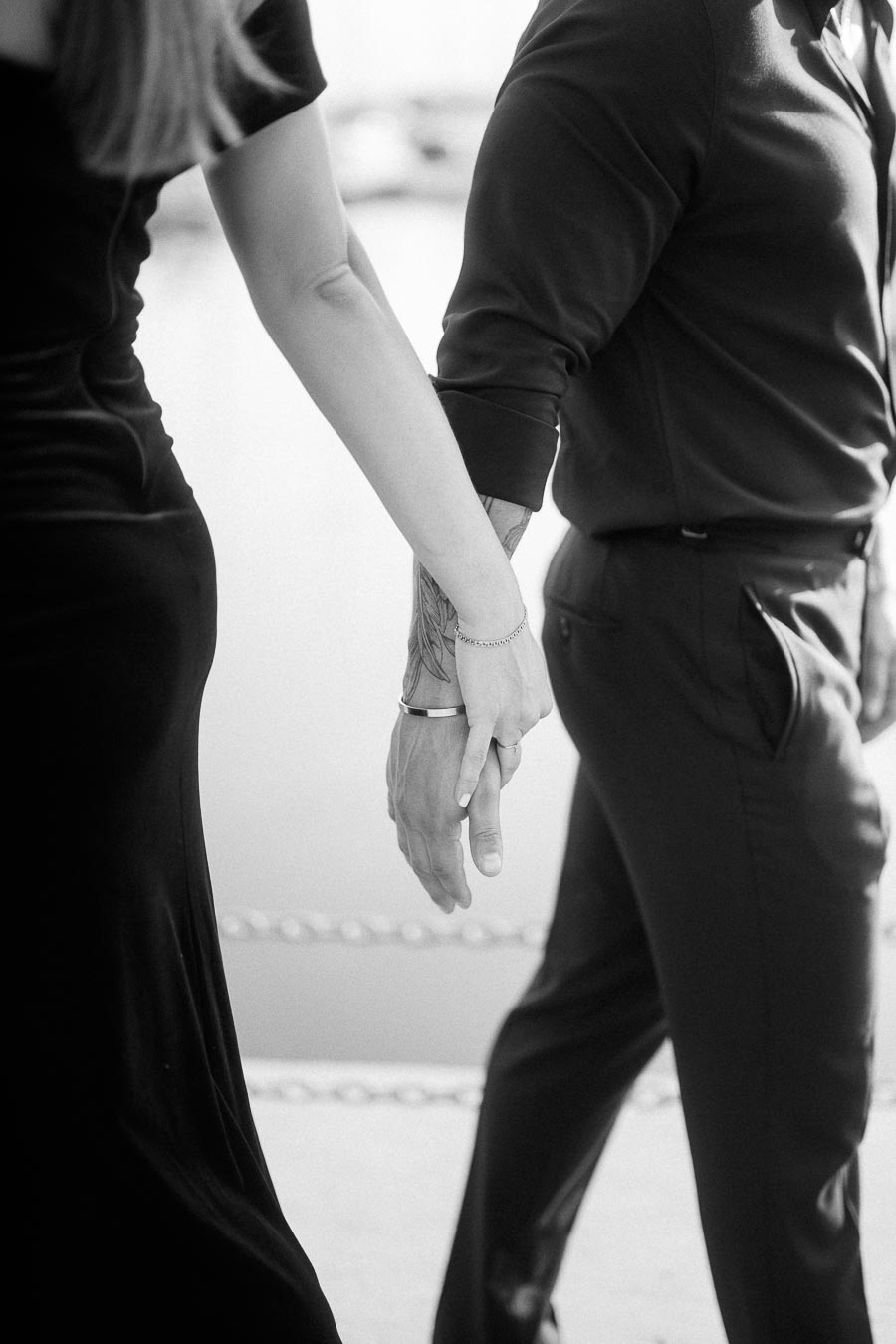 Black-and-white close-up of an engaged couple walking hand in hand near the waterfront.