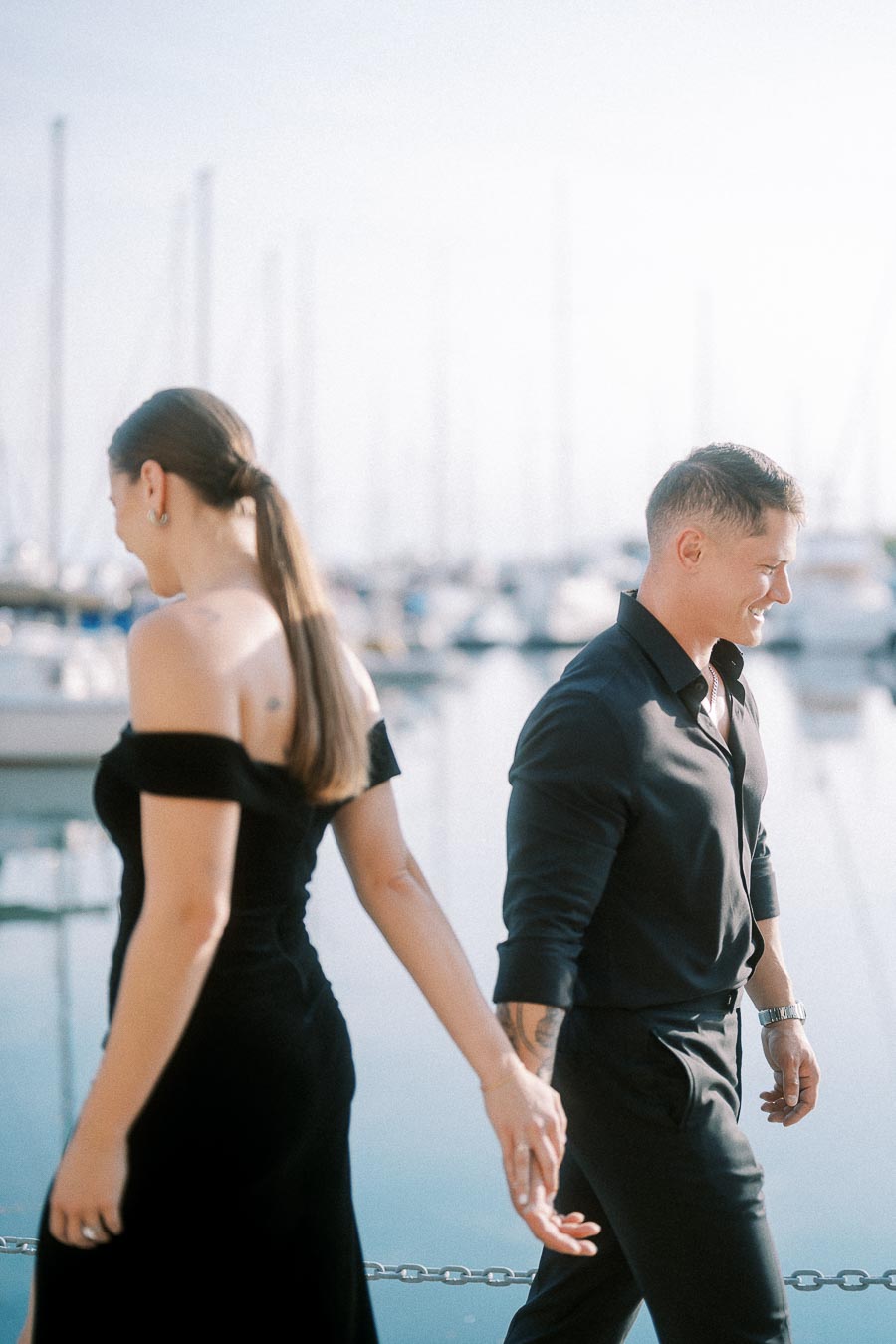 Couple in black outfits holding hands beside a sunlit Toronto marina with sailboats behind them