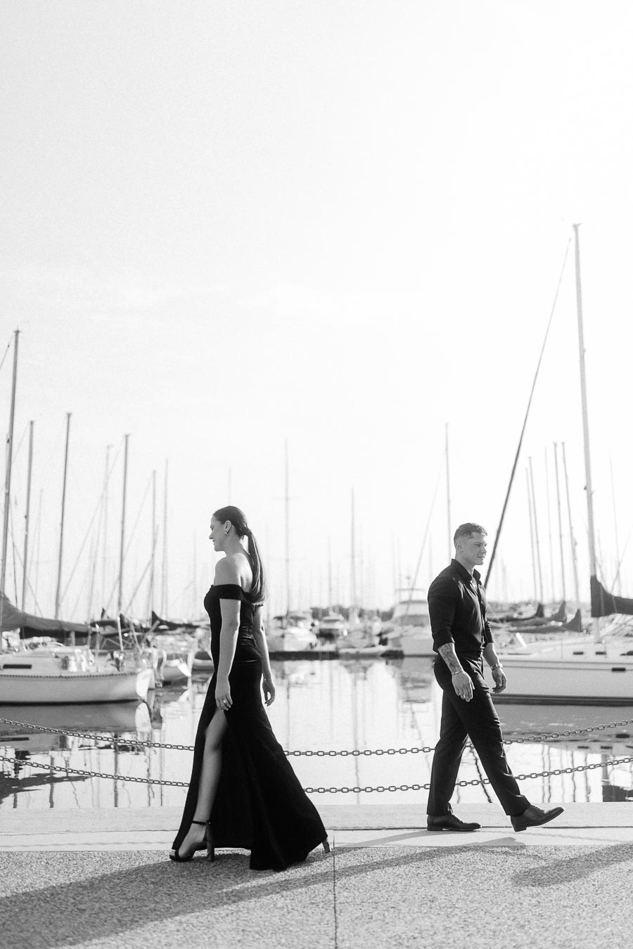 Couple walking separately along a marina promenade with sailboats and reflections behind them.