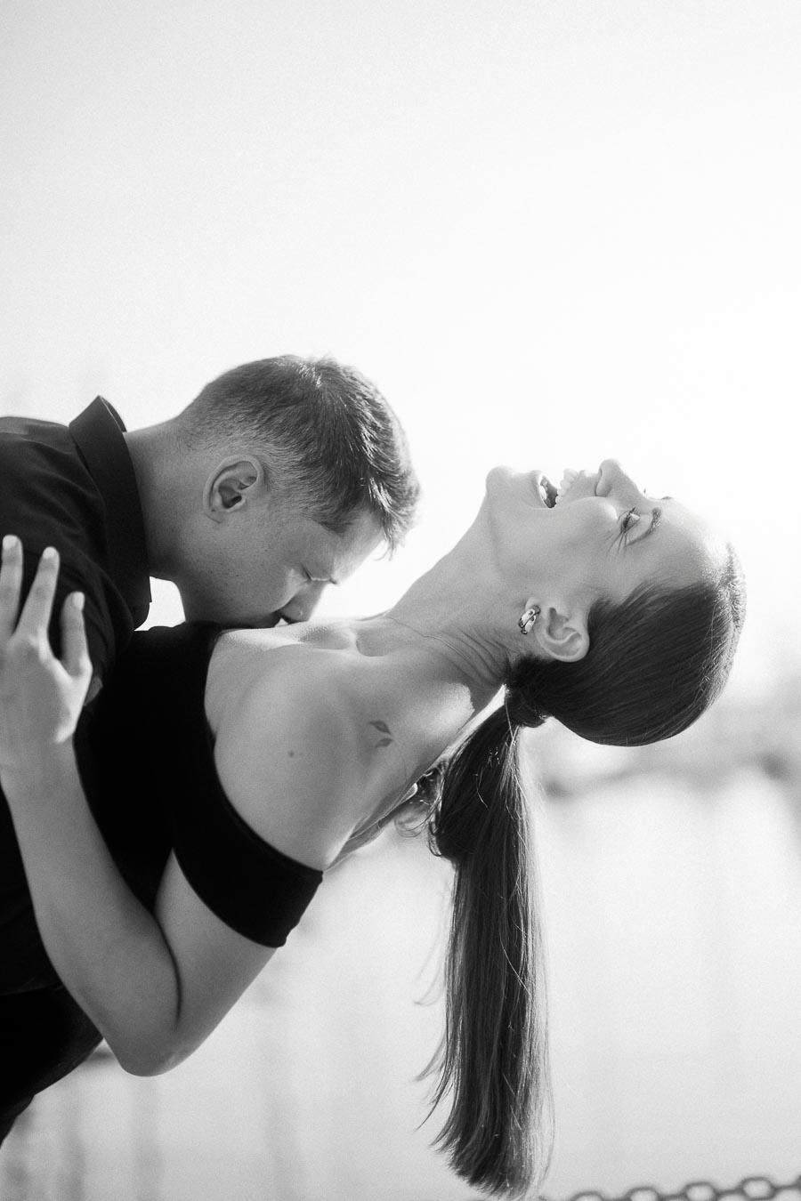 Black and white portrait of groom kissing bride’s shoulder as she laughs in a close embrace.