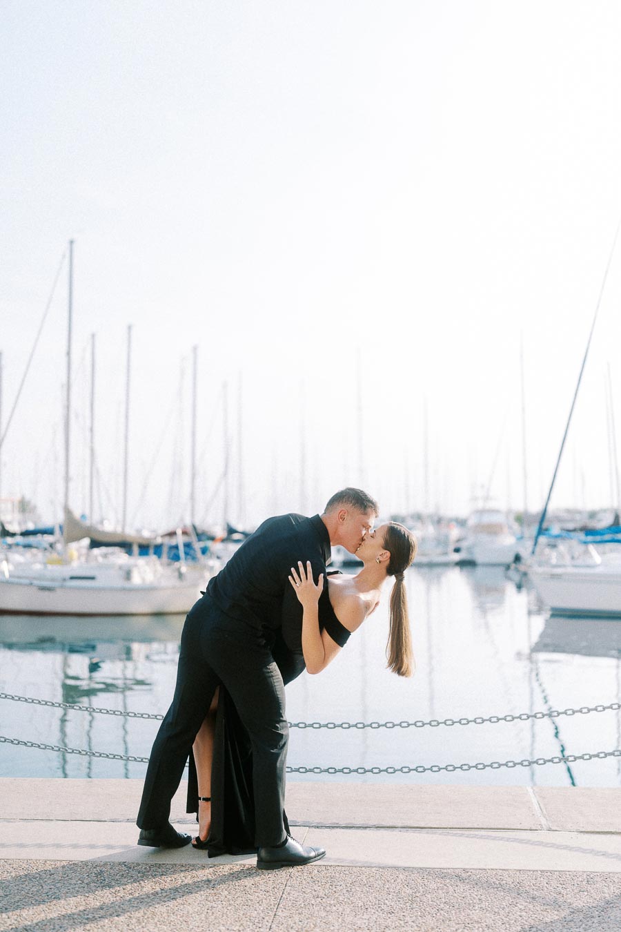 Couple in black formalwear kissing beside a Toronto marina with sailboats reflected in calm water