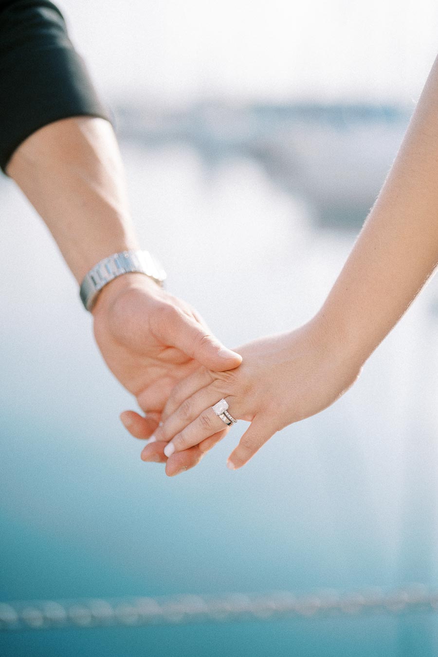 Couple holding hands with an engagement ring visible against a soft waterfront background.