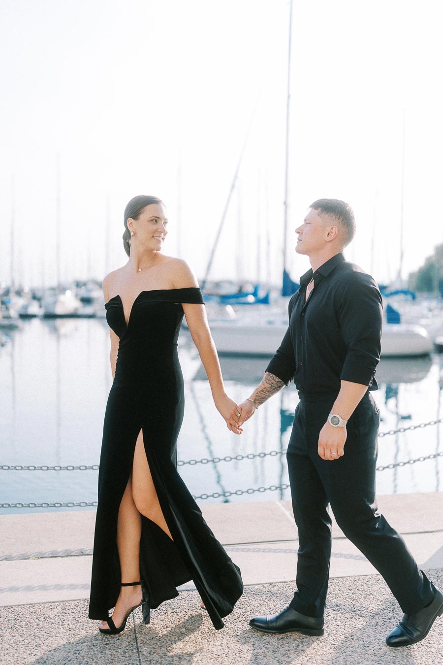 Couple holding hands and walking beside a Toronto marina with sailboats in soft backlight.