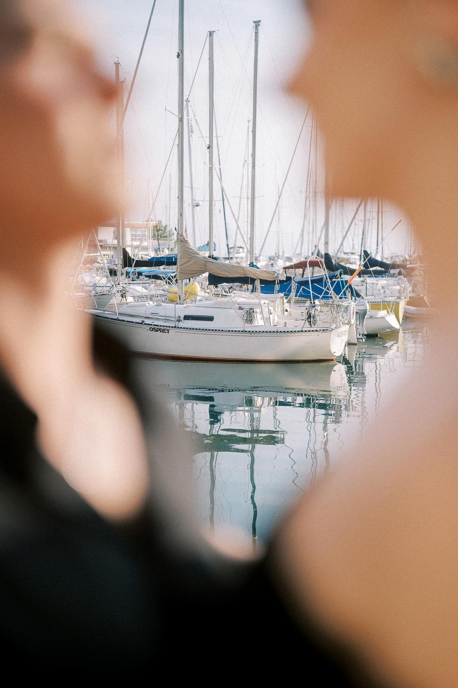 Blurred couple in close foreground with sailboats and marina masts reflected on calm water