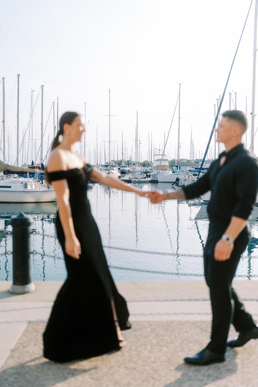 Couple in black attire holding hands beside sailboats at a sunlit Toronto marina.