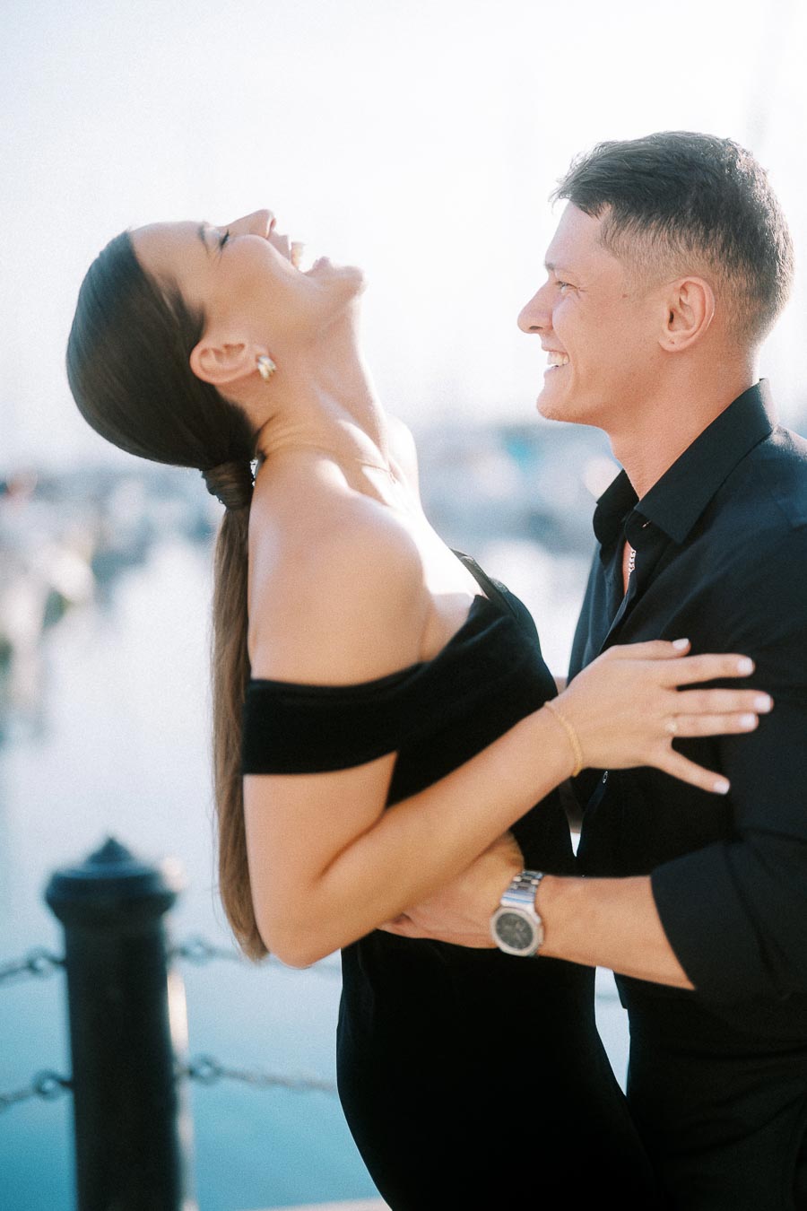Laughing couple in black outfits embracing beside a bright Toronto waterfront marina.