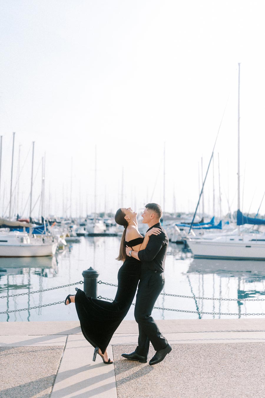 Couple in black formalwear laughing together beside a marina filled with sailboats in Toronto.
