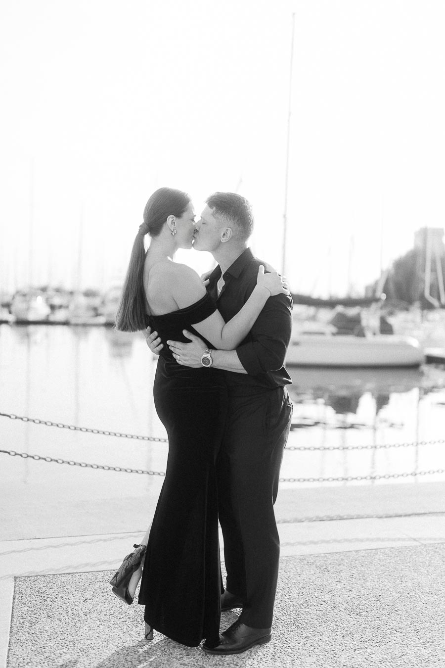 Couple kissing beside a marina, with sailboats and water softly blurred behind them.