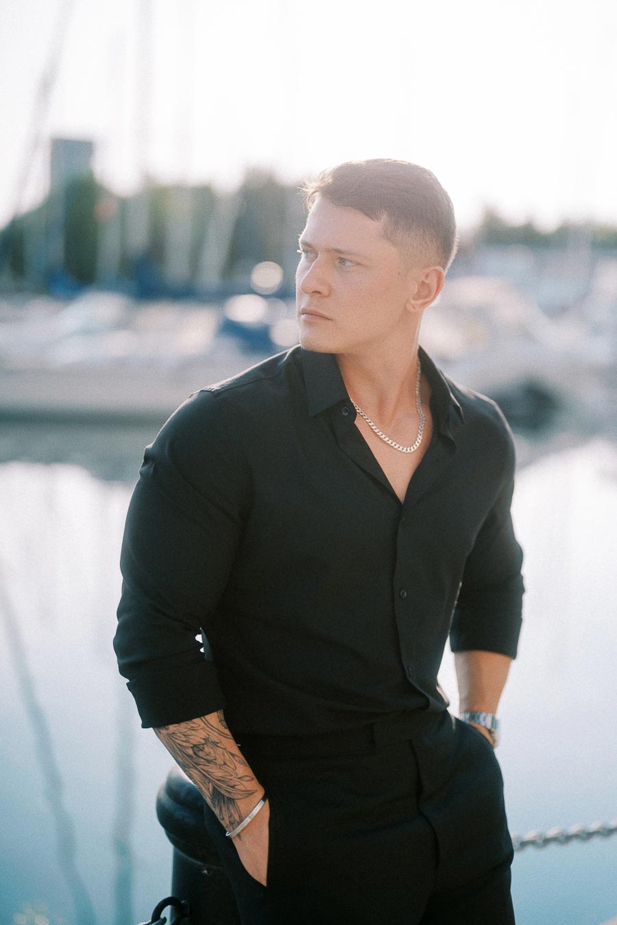 Groom-to-be in black shirt poses beside a marina with blurred sailboats in Toronto.