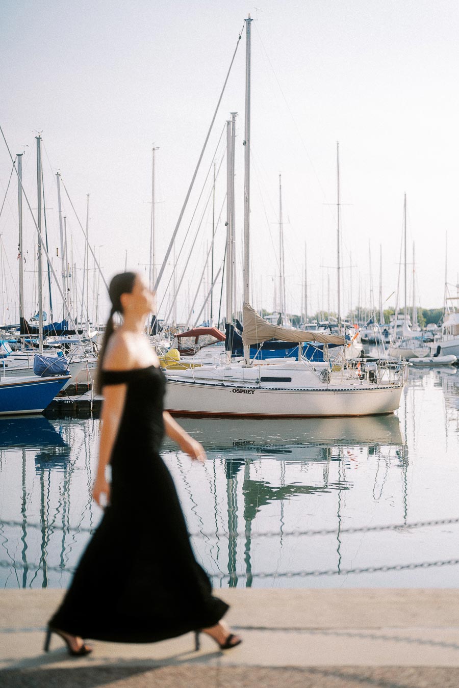 Woman in a black dress walks past moored sailboats at a calm Toronto marina.