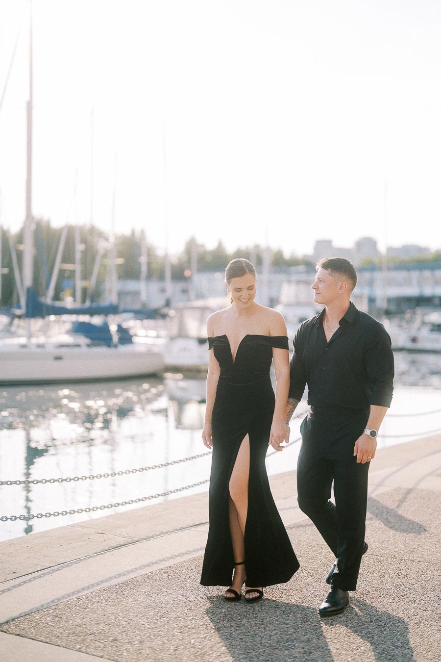 Couple in black formal outfits walking hand in hand beside sailboats at a sunny Toronto marina.