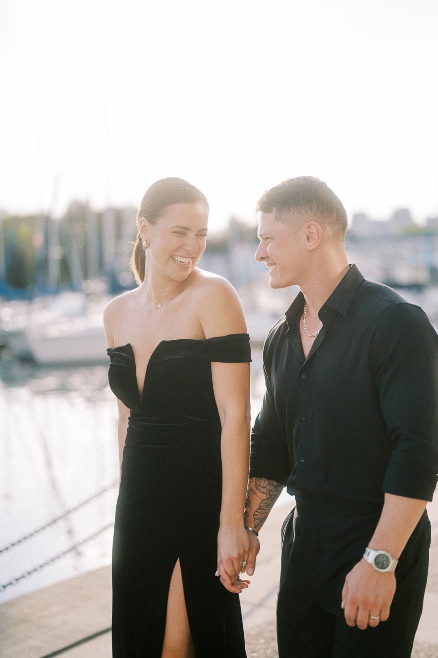 Couple in black outfits holding hands and smiling beside a sunlit Toronto marina.