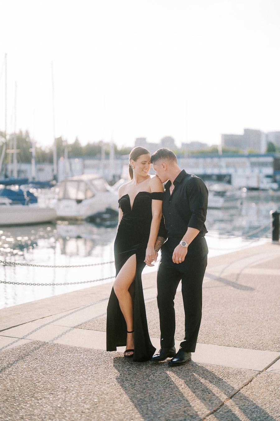 Couple in black formal outfits holding hands beside a sunny Toronto waterfront marina with boats