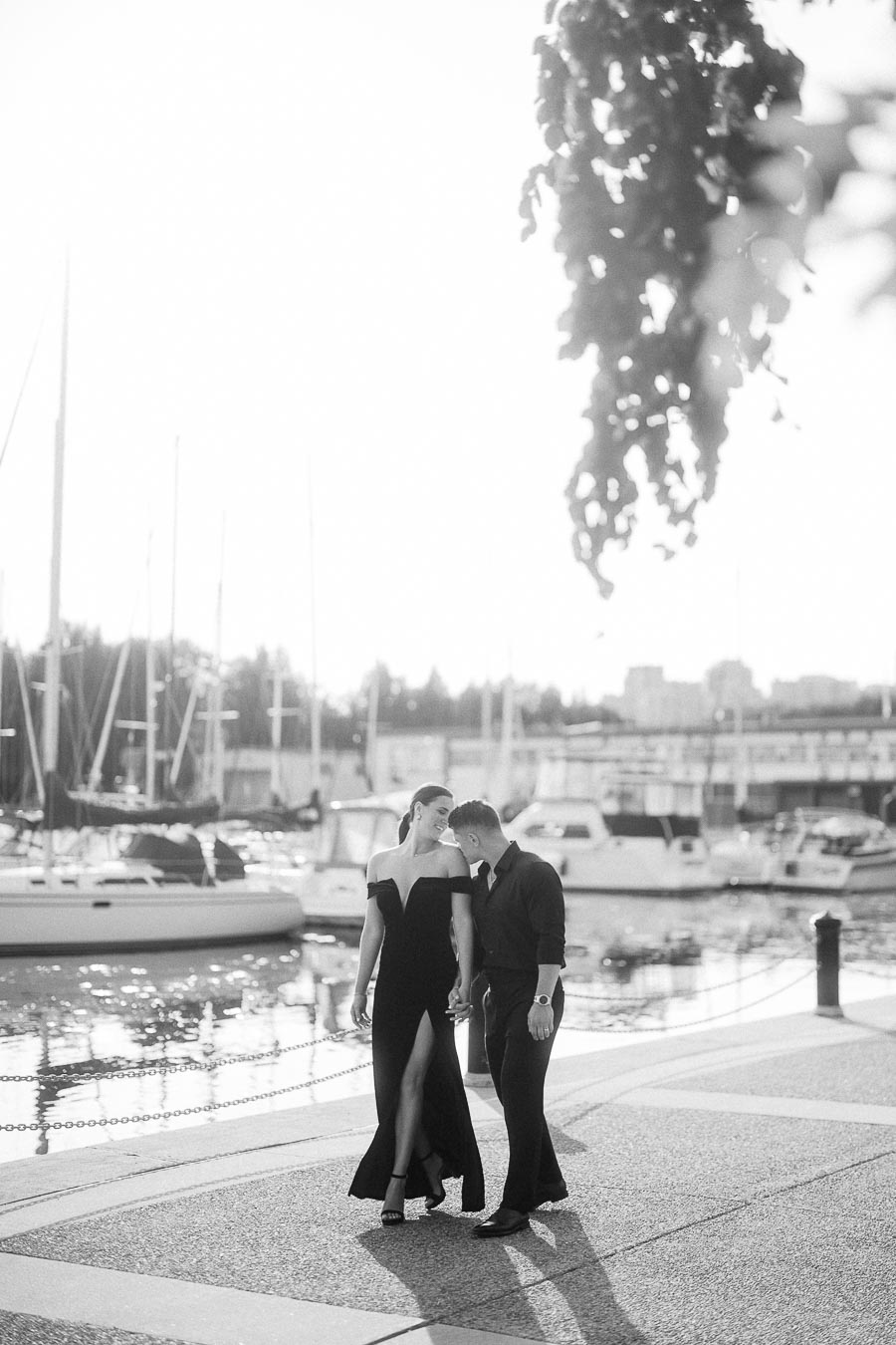 Couple in formal black outfits walking hand in hand beside sailboats at a Toronto waterfront marina.