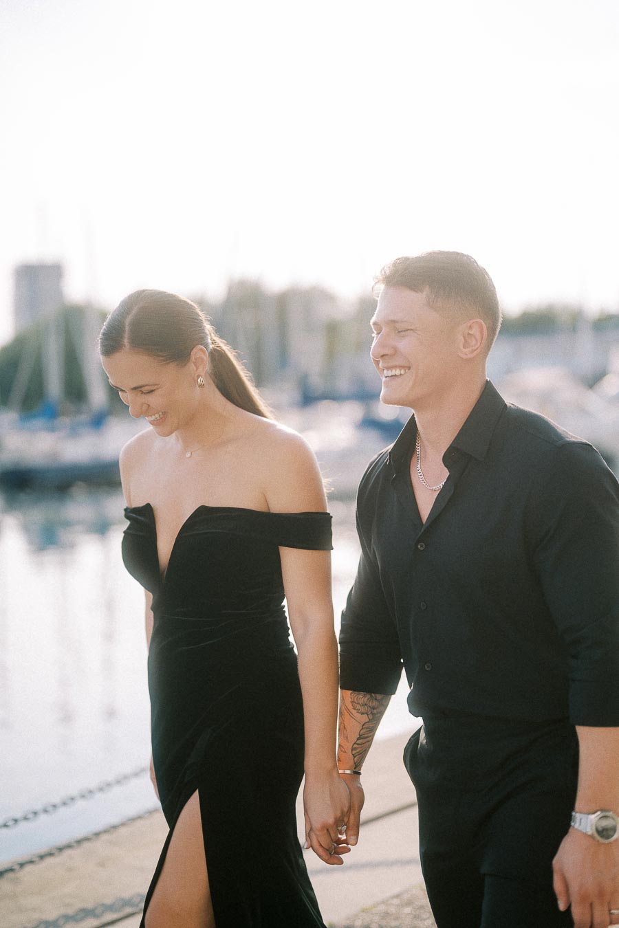 Couple in black outfits holding hands and laughing beside a sunlit Toronto marina.